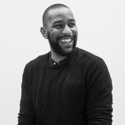 A person with a beard smiles while looking to the side, wearing a dark sweater, against a plain white background.